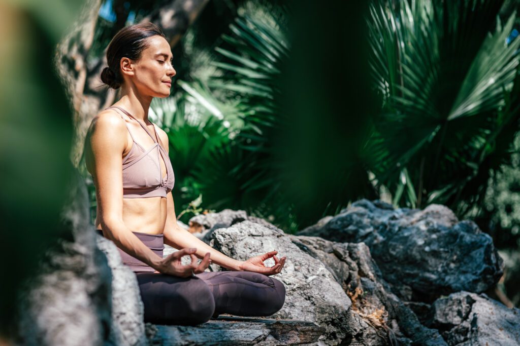 Ganzkörperaufnahme einer kaukasischen Frau, die in einer Easy Seated Pose, Sukhasana, während sie Yoga im Freien in einem Garten praktiziert.