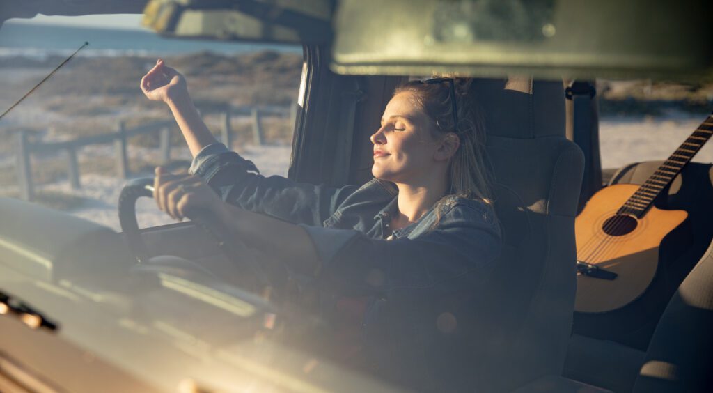 Vorderansicht einer kaukasischen Frau in einem offenen Top-Auto, hält ein Lenkrad, hob ihre Hände und entspannt in der Sonne mit geschlossenen Augen
